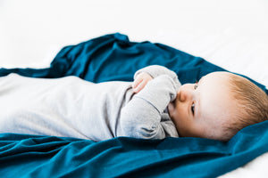 a baby boy cuddled on top of our midnight teal stretch swaddle blanket while wearing our knotted gown in heathered grey