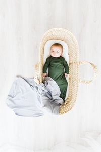 a baby boy in a bassinet wearing our moss green knotted gown with our heathered grey swaddle draped over the bottom of the bassinet