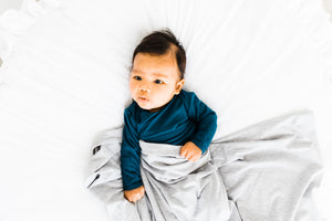 baby boy wearing our knotted baby gown in midnight teal and covered in our heathered grey stretch swaddle blanket