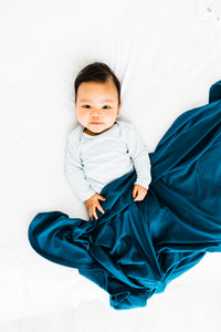 a baby boy covered in our midnight teal stretch swaddle blanket while wearing our kniotted gown in baby blue stripe