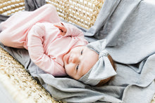 Load image into Gallery viewer, a baby girl laying on our heathered grey swaddle blanket while wearing our knotted gown in light pink and our headband in heathered grey