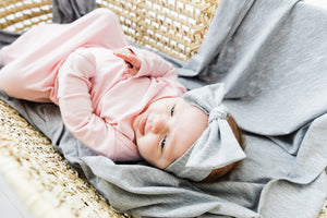 a baby girl laying on our heathered grey swaddle blanket while wearing our knotted gown in light pink and our headband in heathered grey