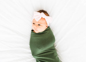 a baby girl wrapped in our moss green bamboo swaddle blanket and wearing our light pink bamboo stretch headband