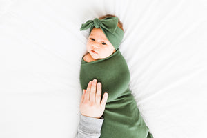 a baby girl wrapped in our moss green bamboo swaddle blanket while also wearing our bamboo stretch headband in moss green